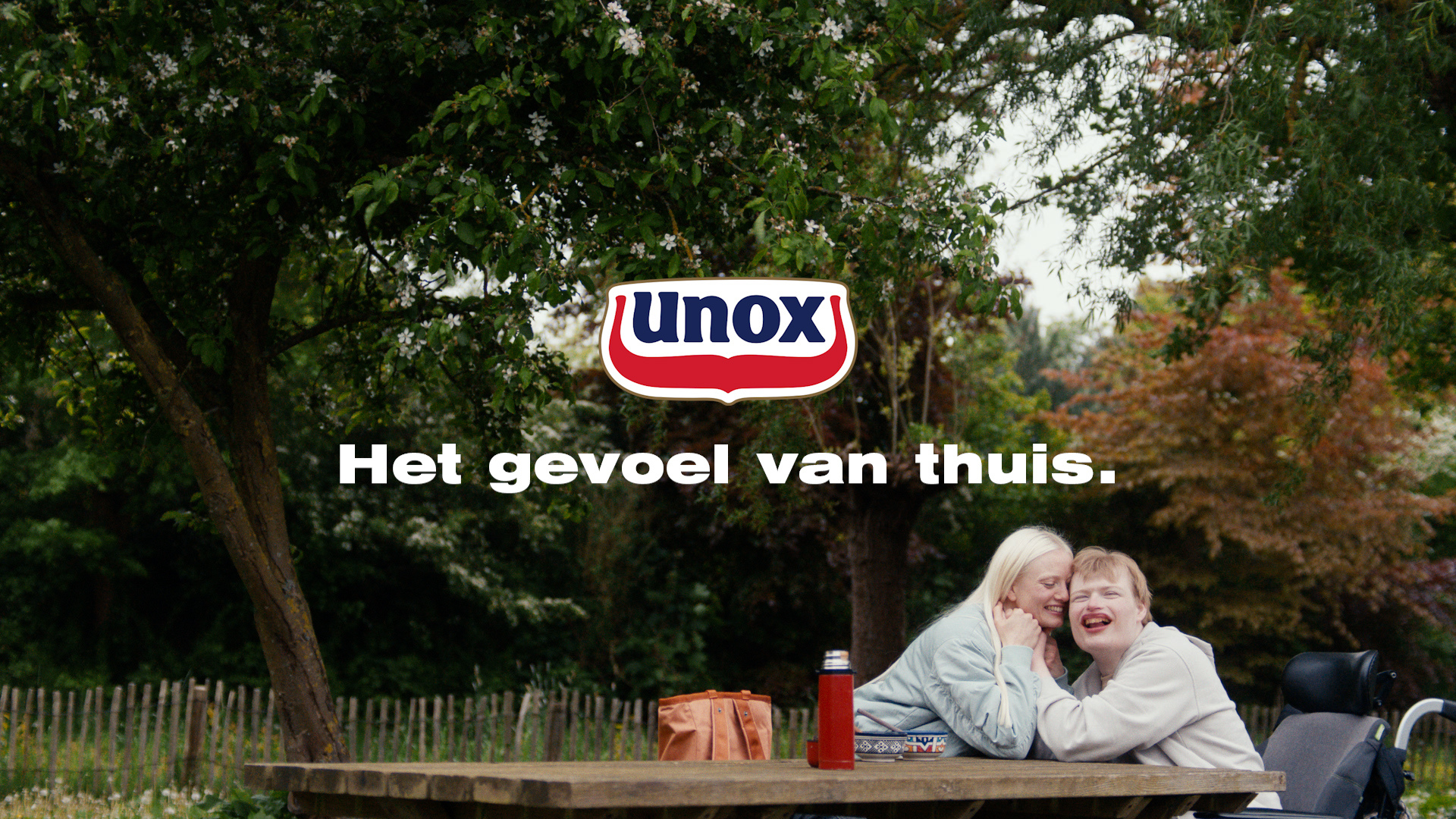 Twee mensen zitten aan een picknicktafel in een park. De vrouw in de rolstoel lacht terwijl de andere vrouw naar haar toe leunt om haar te knuffelen.