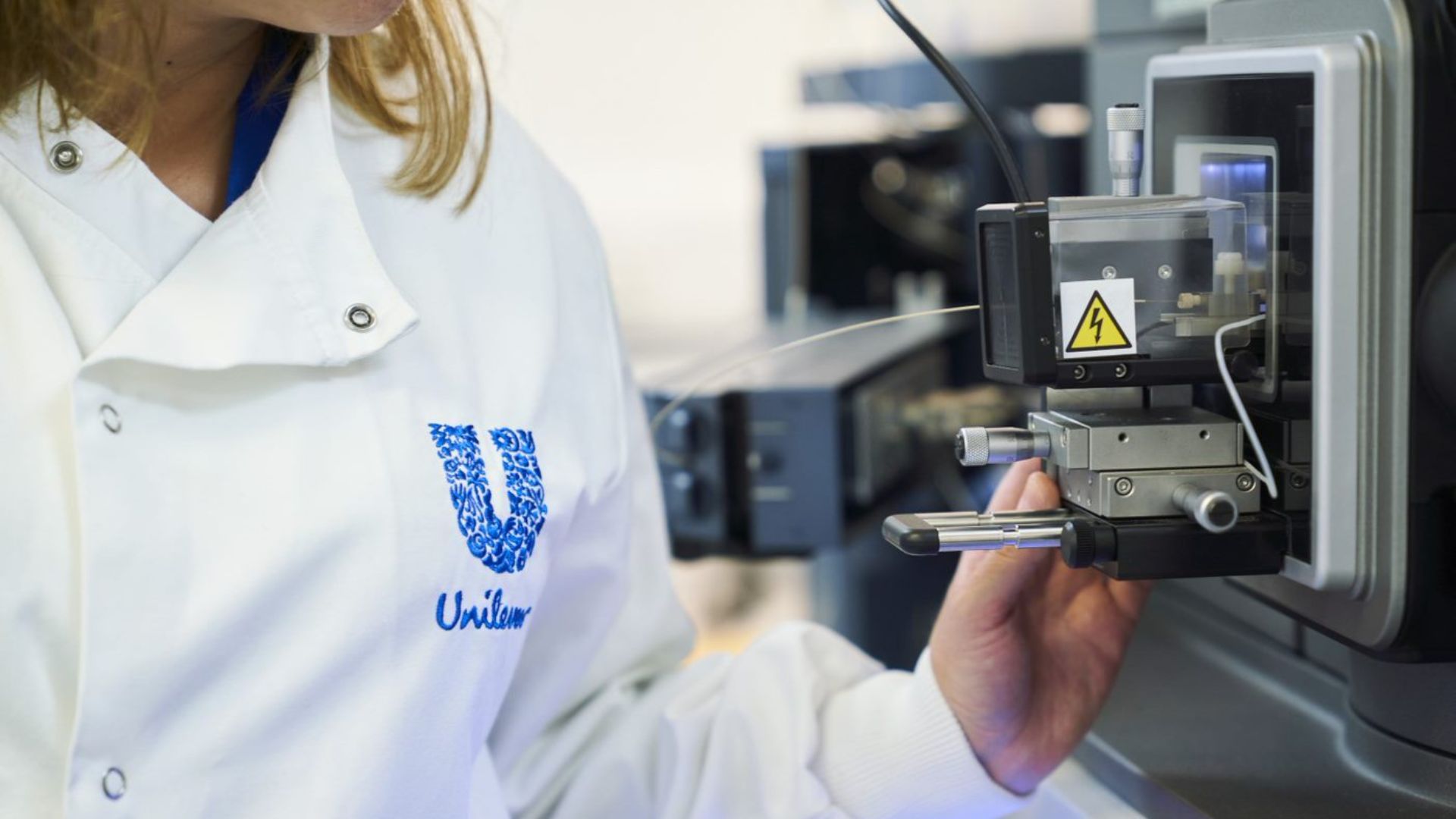 Een close-upfoto van een wetenschapper in een witte laboratoriumjas van Unilever, die door een grote digitale microscoop kijkt.