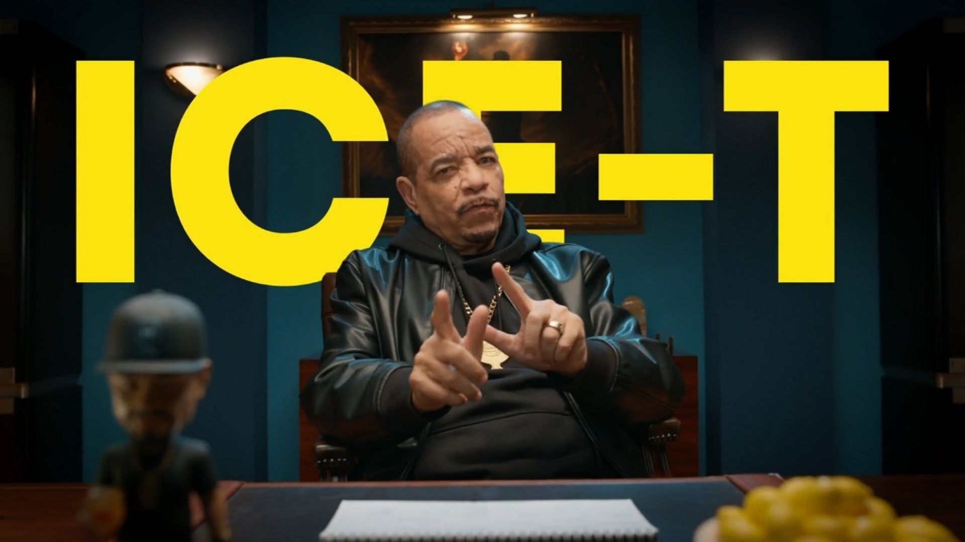 Persoon die aan een bureau zit en met beide handen gebaart, met grote gele tekst "ICE-T" op de achtergrond.