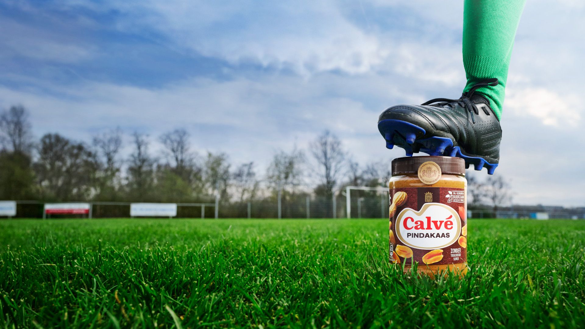 Een voet met een voetbalschoen rust op een pot Calvé pindakaas op een grasveld.